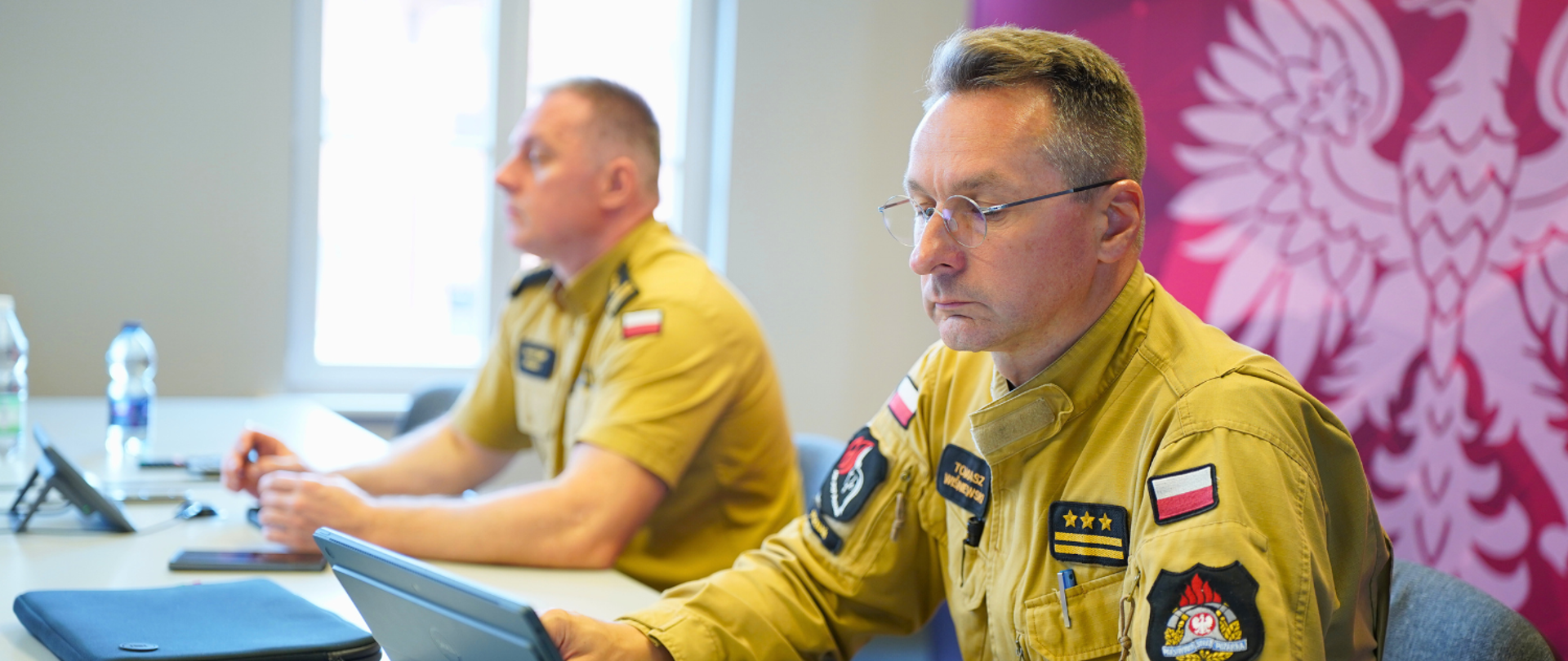 Naczelnicy wydziałów oraz Panowie Komendanci uczestniczą w wideokonferencji w sali prezydialnej komendy, siedząc za stołami, wpatrując się w obraz wyświetlany na telewizorze. 