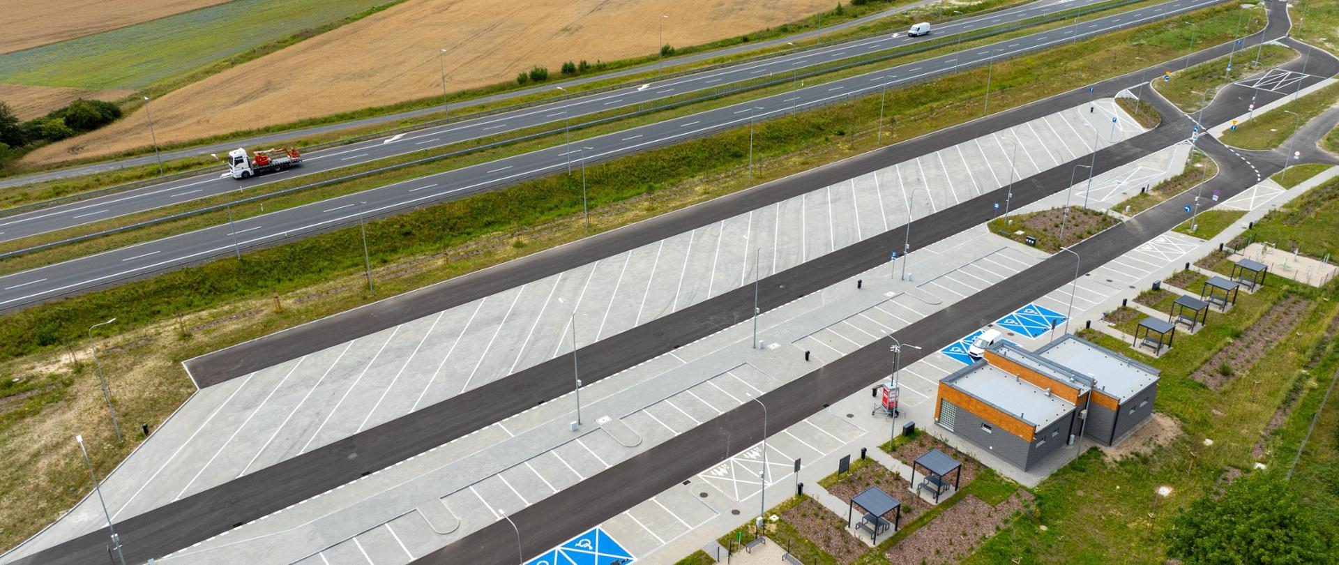 To zdjęcie przedstawia fragment drogi ekspresowej widziany z lotu ptaka. Droga ma wiele pasów ruchu w obu kierunkach, oddzielonych trawiastym pasem w środku. Po prawej stronie zdjęcia znajduje się zjazd prowadzący do Miejsca Obsługi Podróżnych, parkingu, na którym zaparkowane są samochody. Wokół drogi rozciągają się pola uprawne o różnych odcieniach zieleni i brązu, wskazujące na różne rodzaje roślinności. 