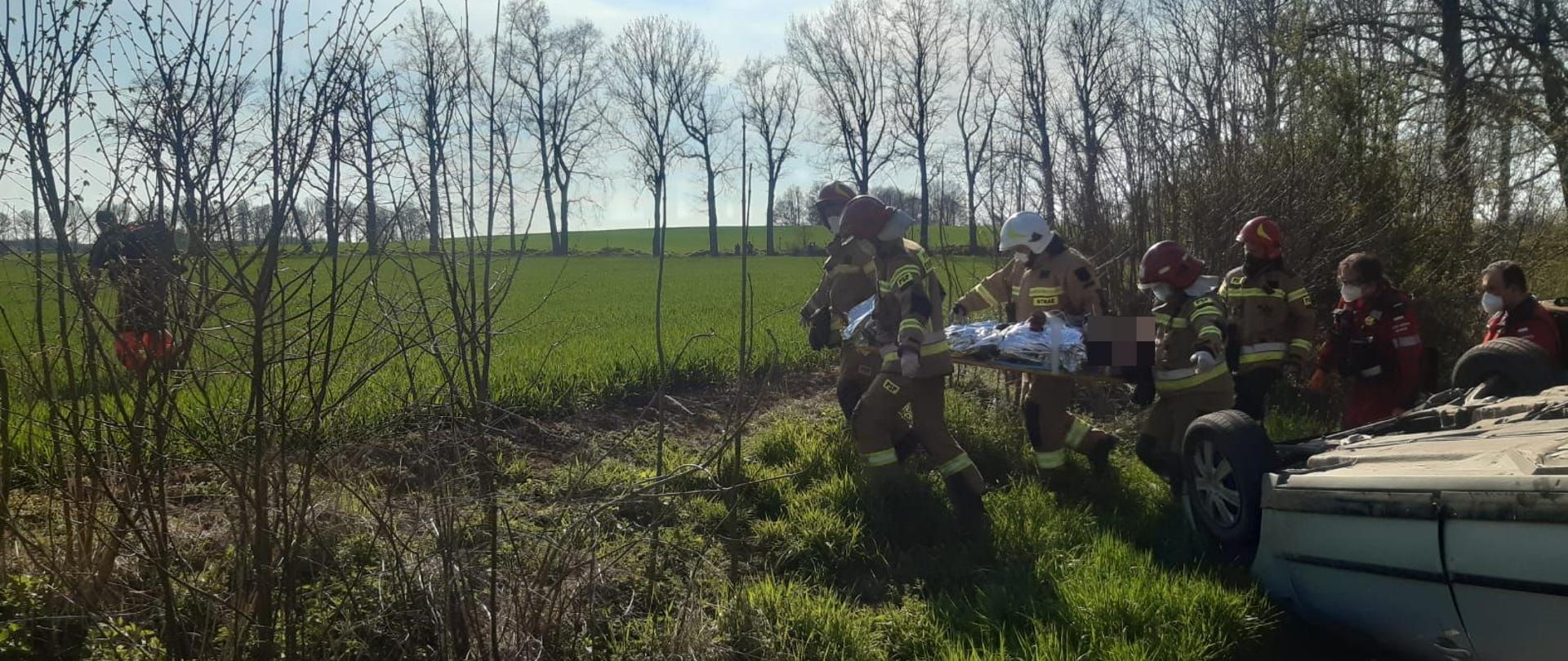 Zdjęcie przedstawia strażaków podczas pomocy w transporcie osoby poszkodowanej na desce w kierunku śmigłowca Lotniczego Pogotowia Ratunkowego. Za strażakami widoczna obsada śmigłowca a koło nich samochód na dachu.