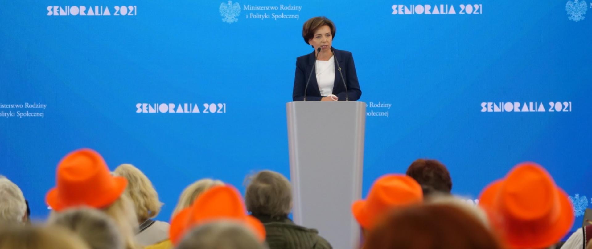 Minister Maląg przemawia do zgromadzonych stojąc na mównicy. W tle niebieski baner z białym napisem Senioralia 2021 oraz z logo Ministerstwa Rodziny. Na pierwszym planie sylwetki siedzących ludzi. Widok pleców. Niektórzy mają na głowach pomarańczowe kapelusze.