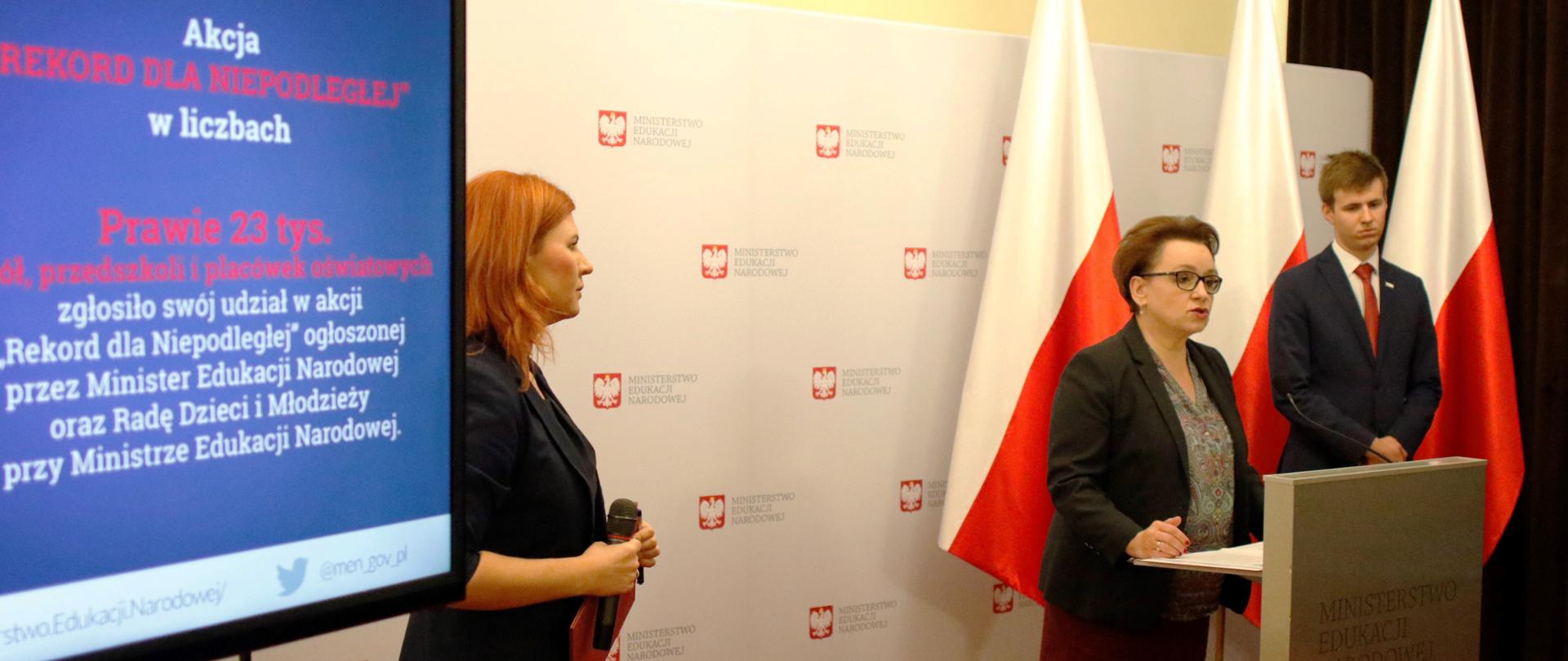 Konferencja prasowa Minister Edukacji Narodowej Anny Zalewskiej. W tle monitor z wyświetlaną planszą o akcji Rekord dla Niepodległej. W tle biało czerwone-flagi