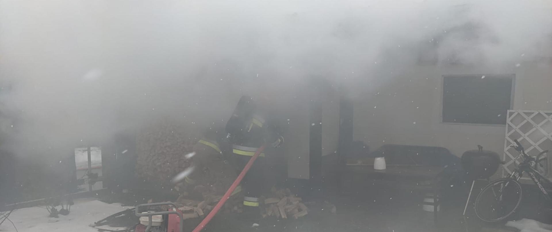 Zdjęcie przedstawia dwóch strażaków (obecnych jeszcze na zewnątrz) wyposażonych w sprzęt ochrony układu oddechowego i linię gaśniczą, podejmujących działania gaśnicze podczas pożaru budynku garażowego. Na obrazie widoczny jest dym wydobywający się z obiektu