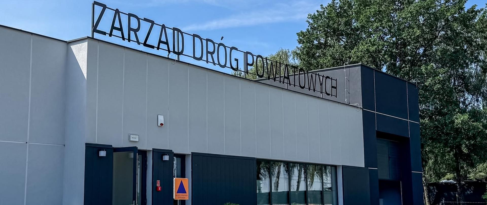 Budynek Zarządu Dróg Powiatowych widziany z zewnątrz, z zielonymi roślinami na pierwszym planie i widocznym napisem na elewacji