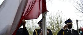 Trzej strażacy z pocztu flagowego wciągają flagę Rzeczypospolitej Polskiej na maszt. Jeden ze strażaków podtrzymuje flagę, drugi wciąga ją za pomocą linki na maszt, a trzeci oddaje honory. W tle widać strażaków zmiany przyjmującej służbę oraz ogrodzenie i zarośla.