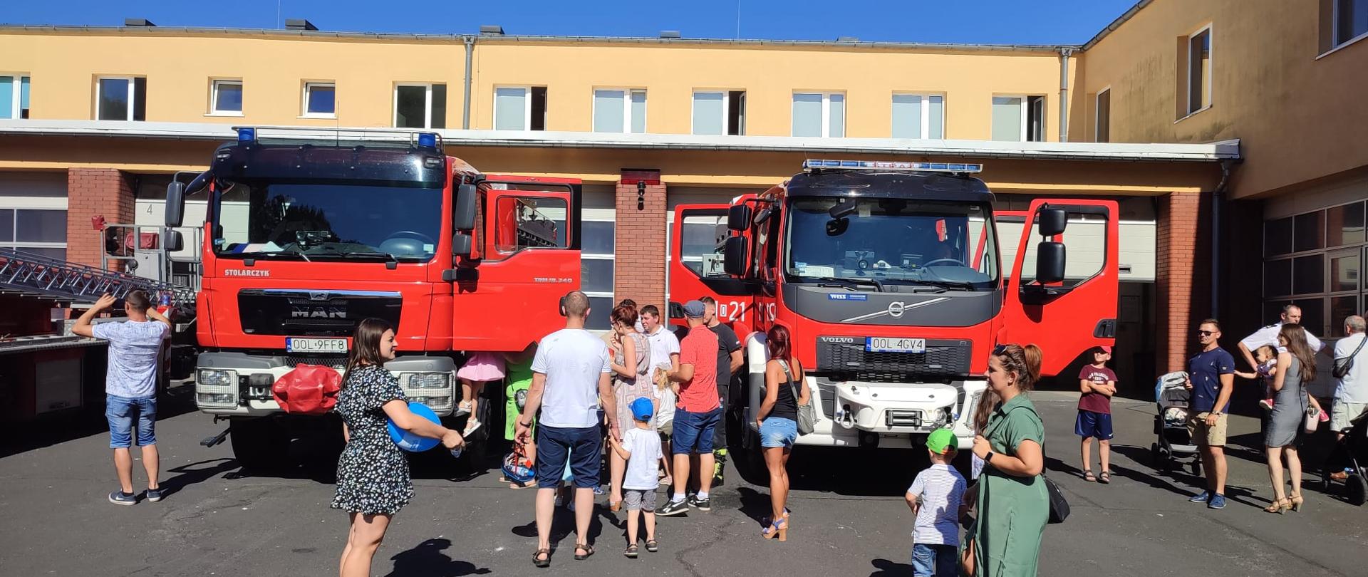 Na zdjęciu widać rodziny oglądające samochody pożarnicze na tle dwóch samochodów pożarniczych stojących na placu przed garażami.
