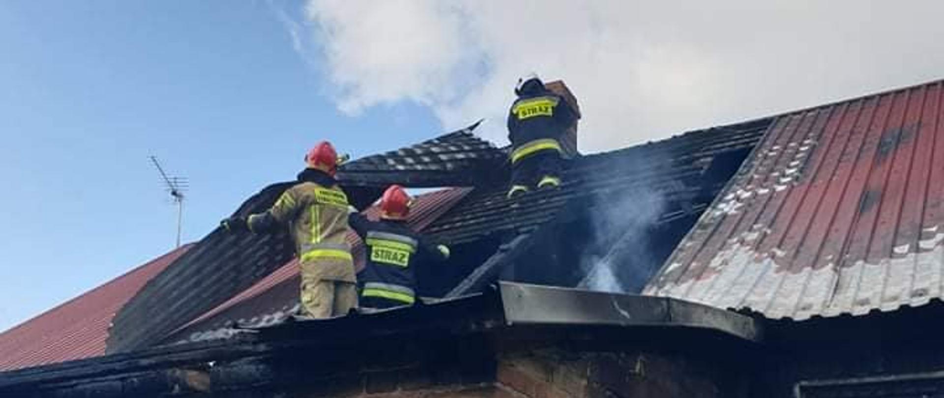 Strażacy pracujący na dachu przy demontażu nadpalonej konstrukcji dachu. Futryny drzwi i okien są nadpalone. Na blachodachówce widać ślady działania ognia.