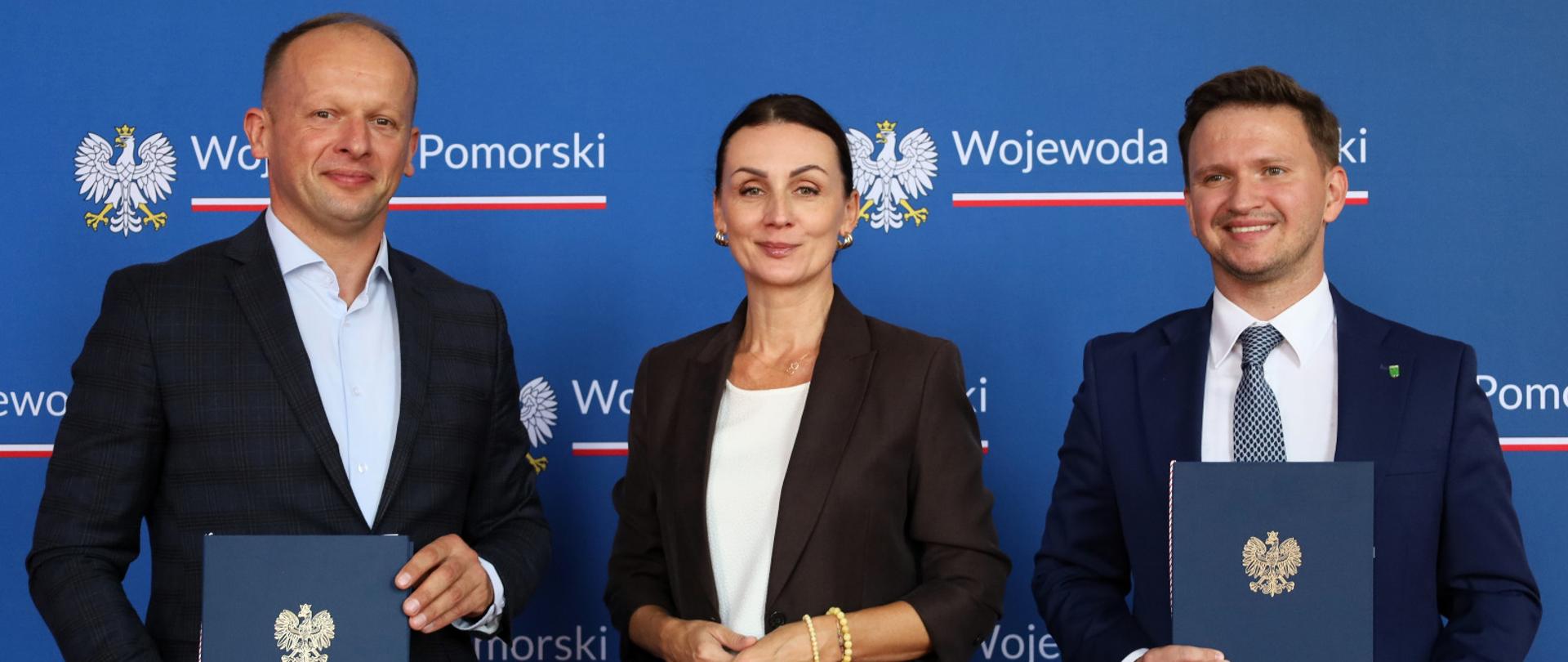 trzy osoby stoją na tle tablicy z napisem wojewoda pomorski