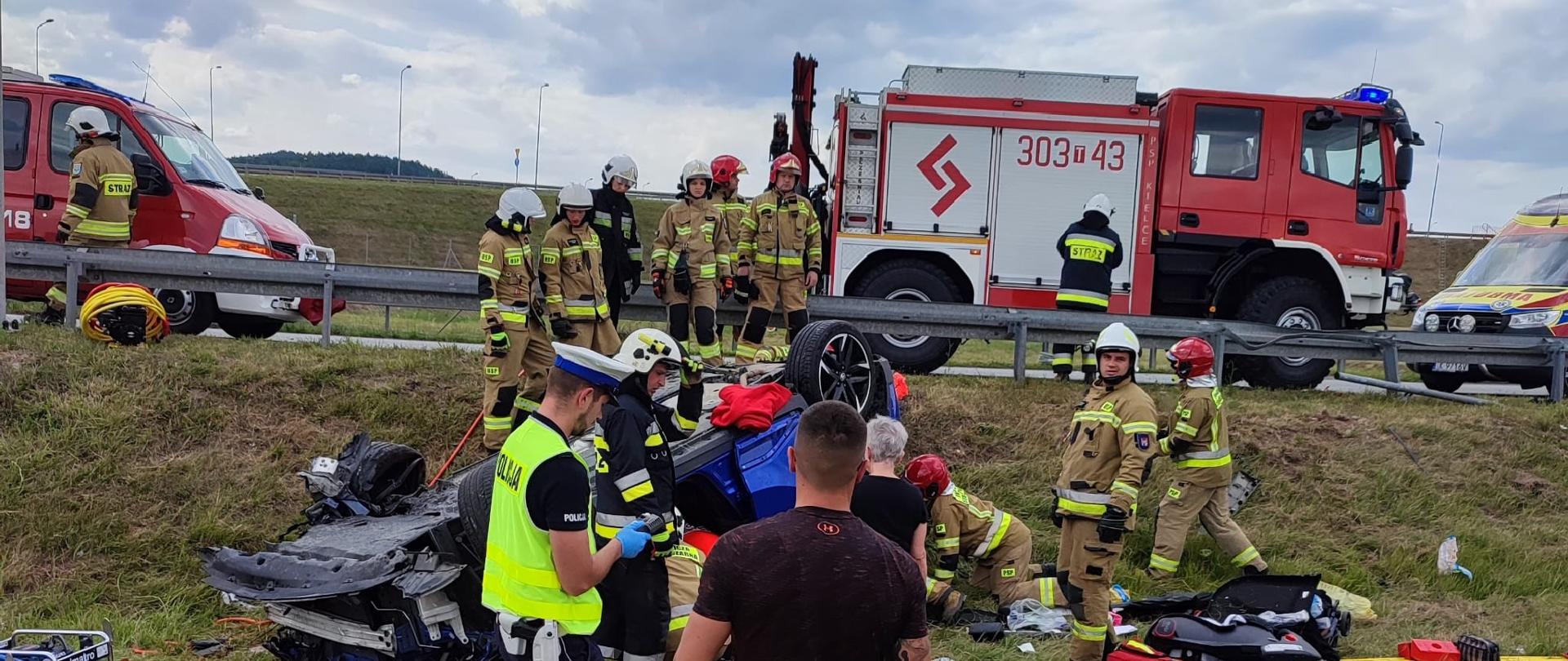 Zdjęcie przedstawia miejsce działań. Na pierwszym planie pobocze, na którym leży na dachu samochód. Wokół niego strażacy , ratownicy medyczni oraz funkcjonariusze Policji. Na ziemi porozrzucane bagaże, elementy zniszczonego auta oraz sprzęt ratowniczy. Powyżej, na jezdni stoją dwa auta pożarnicze oraz ambulans.