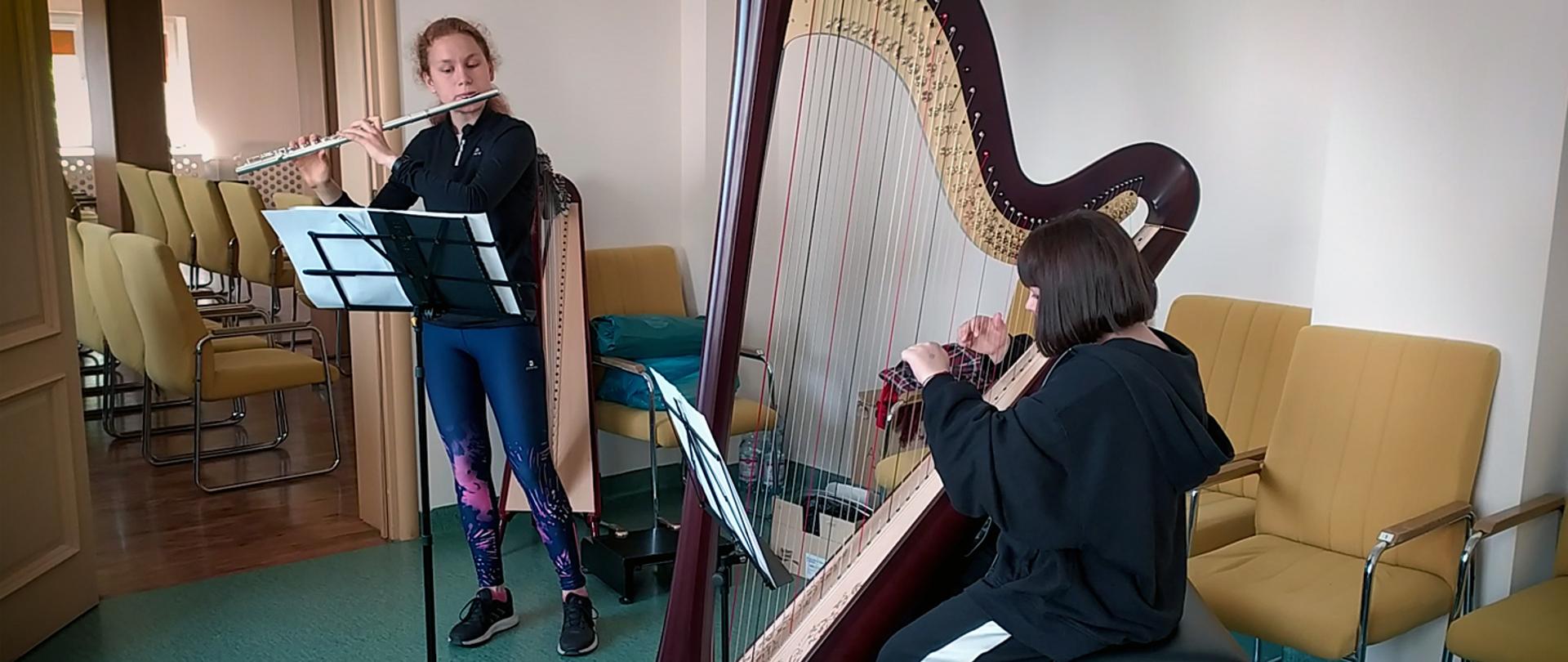 Dwie dziewczyny grają na instrumentach w sali prób. Dziewczyna z lewej stojąc gra na flecie, dziewczyna z prawej, siedząc gra na harfie.