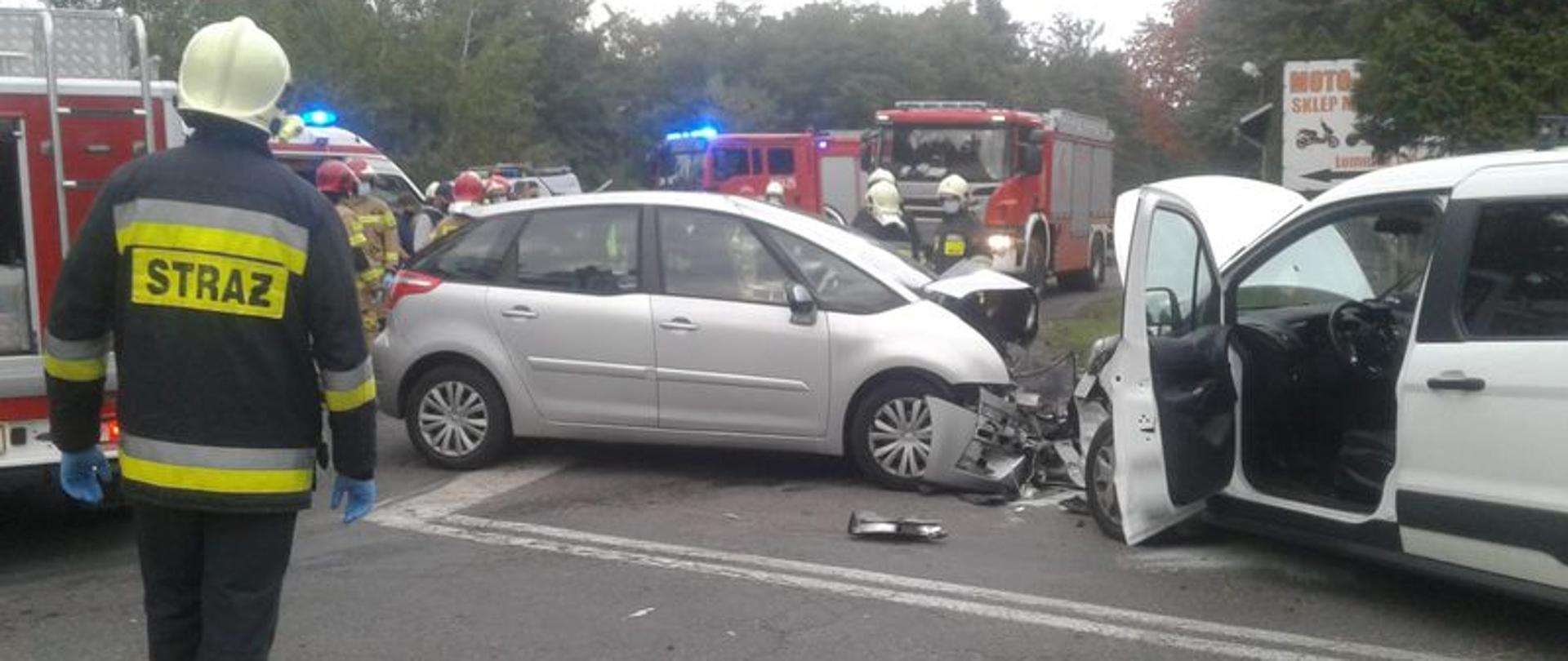 Dzień. Dwa uszkodzone samochody osobowe mają rozbite przody. W tle wozy strażackie, kartka pogotowia ratunkowego oraz strażacy.