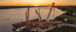 Dwa duże dźwigi podnoszą długi cylindryczny element przemysłowy nad brzegiem rzeki o zachodzie słońca.