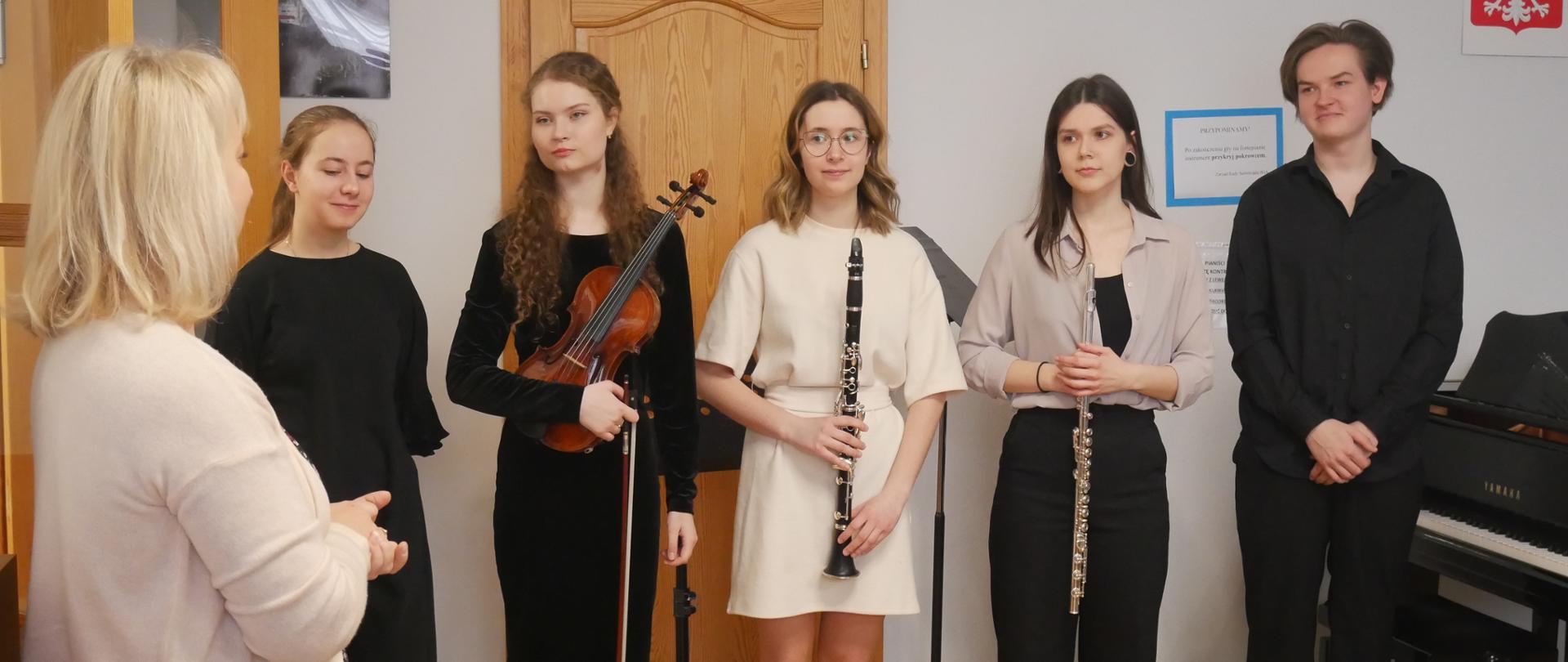 Wykonawcy kłaniają się publiczności. Od lewej strony stoją: solistka Ania, skrzypaczka Roksana, klarnecistka Sonia i waltornista Bartek, który wystąpił w roli pianisty. Przed muzykami stoi pani Ania, nauczycielka ze szkoły specjalnej i dziękuje za występ. 