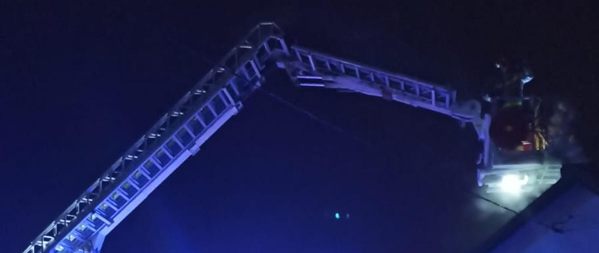 Na pierwszym planie widnieje metalowe ogrodzenie posesji. Za nim przedstawiony jest parterowy budynek mieszkalny. Przy budynku znajduje się specjalny samochód z podnośnikiem hydraulicznym. Pojazd ma rozłożony park drabinowy a w jego koszu zarysowana jest sylwetka strażaka przy kominie budynku. Zdjęcie zrobione jest w porze nocnej, wokół panuje mrok z odblaskami niebieskich świateł alarmowych włączonych w
samochodzie ratowniczo-gaśniczym. Na ziemi zalega biała warstwa śniegu.
