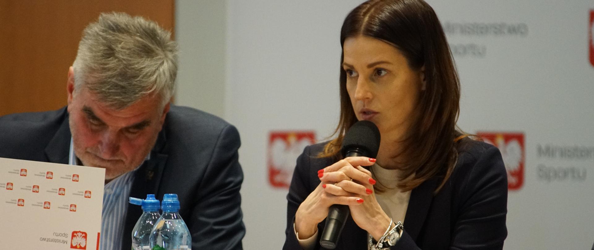 Minister Sportu Danuta Dmowska-Andrzejuk siedzi przy stole prezydialnym i przemawia. W tle ścianka z logo Ministerstwa Sportu