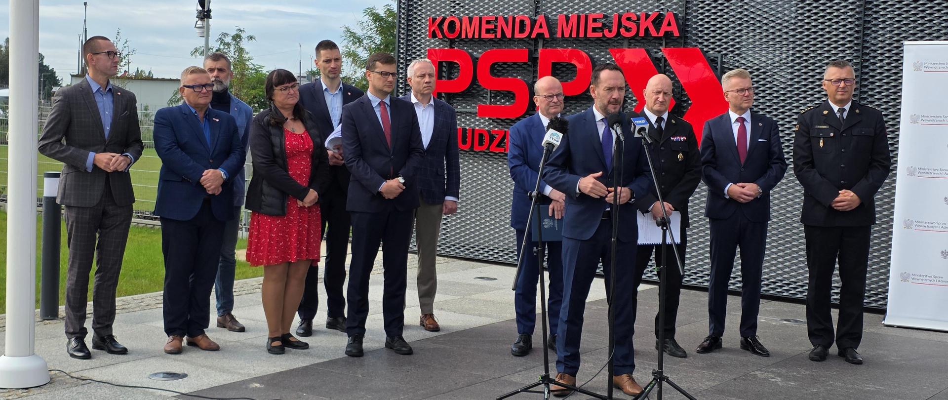 Grupa osób stojących przed budynkiem podczas konferencji. Mężczyzna przy mikrofonie przemawia do zebranych.