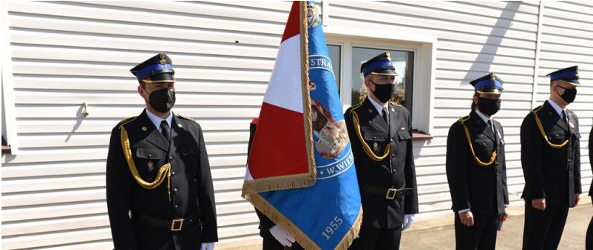 Zdjęcie przedstawia pięciu funkcjonariuszy Państwowej Straży Pożarnej w umundurowaniu wyjściowym w tym trzech wchodzących w skład Pocztu Sztandarowego w białych rękawiczkach. Drugi funkcjonariusz od lewej trzyma sztandar Komendy Powiatowej Państwowej Straży Pożarnej w Wieruszowie. Wszyscy stoją na tle budynku Jednostki Ratowniczo - Gaśniczej Powiatowej Państwowej Straży Pożarnej w Wieruszowie.