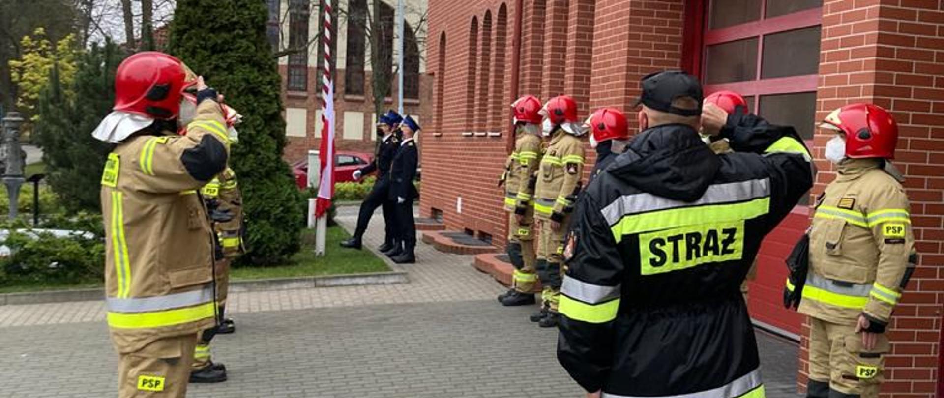 Przed bramami garażowymi JRG Sopot stoja strażacy w dwóch szeregach na przeciw siebie podczas zmainy służby. w tyle trwa wciąganie flagi na maszt p[rzez poczet flagowy