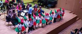 Około 20 dzieci w tych samych kolorowych strojach stoją w rzędzie i śpiewają, za nimi stoi mężczyzna i dyryguje, gra orkiestra.