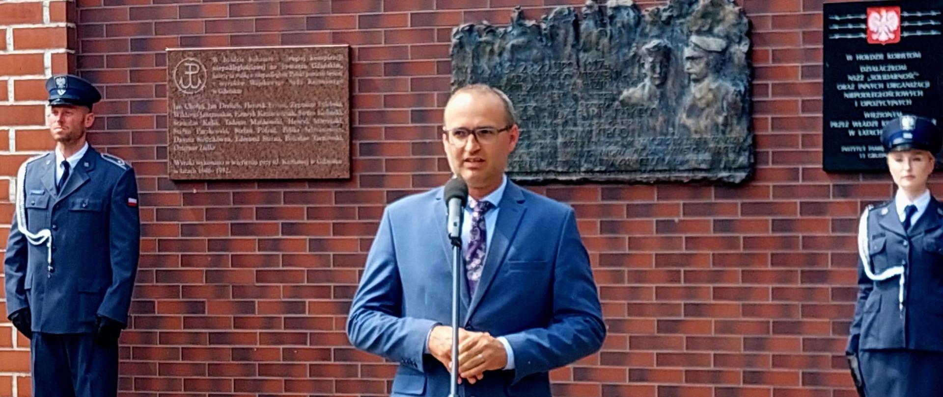 mężczyzna stoi przed mikrofonem i przemawia. W tle mury aresztu śledczego i tablica pamiątkowa poświęcona marszałkowi Piłsudskiemu