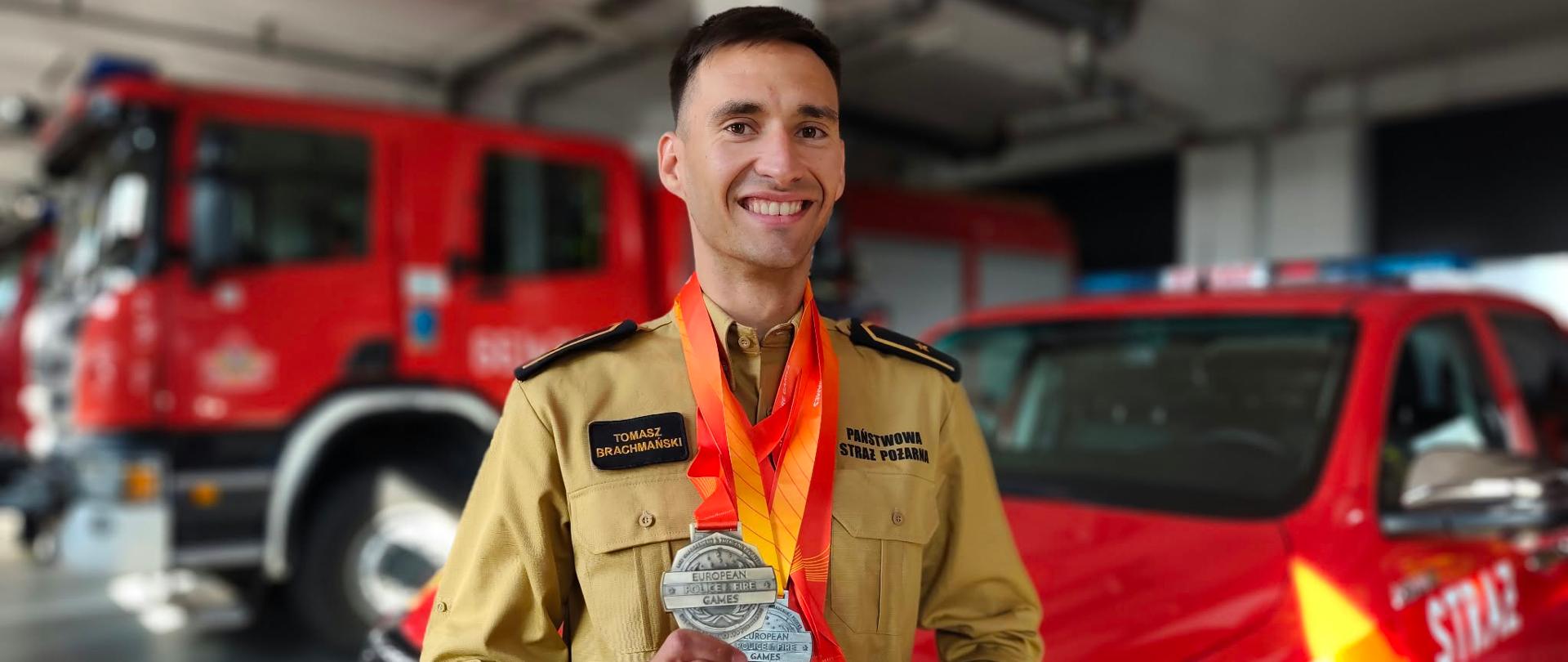 Strażak Państwowej Straży Pożarnej Tomasz Brachmański prezentuje zdobyte medale podczas European Police & Fire Games na tle pojazdów pożarniczych w garażu jednostki.