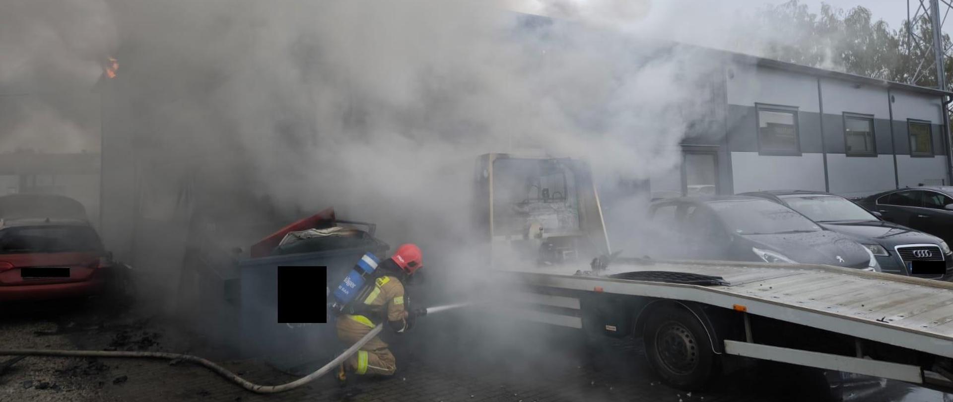 Z kłębów dymu wyłania się budynek hali. Obok stoi zaparkowana, częściowo nadpalona autolaweta samochodowa. Tuż obok niej znajduje się strażak z wężem pożarniczym.