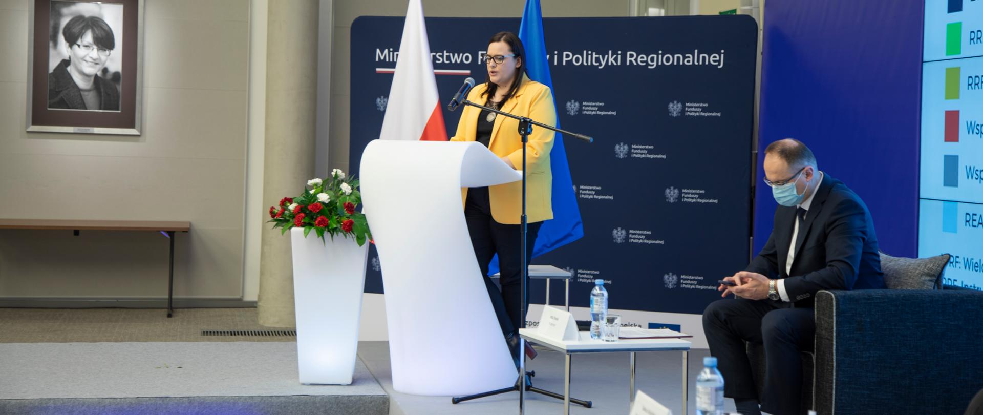 W sali konferencyjnej przy białej mównicy stoi wiceminister Małgorzata Jarosińska-Jedynak. Z lewej strony biało-czerwone kwiaty. Na ścianie portret Grażyny Gęsickiej. Z prawej strony w fotelu siedzi mężczyzna. Z tyłu flagi PL i UE oraz ścianka MFiPR.