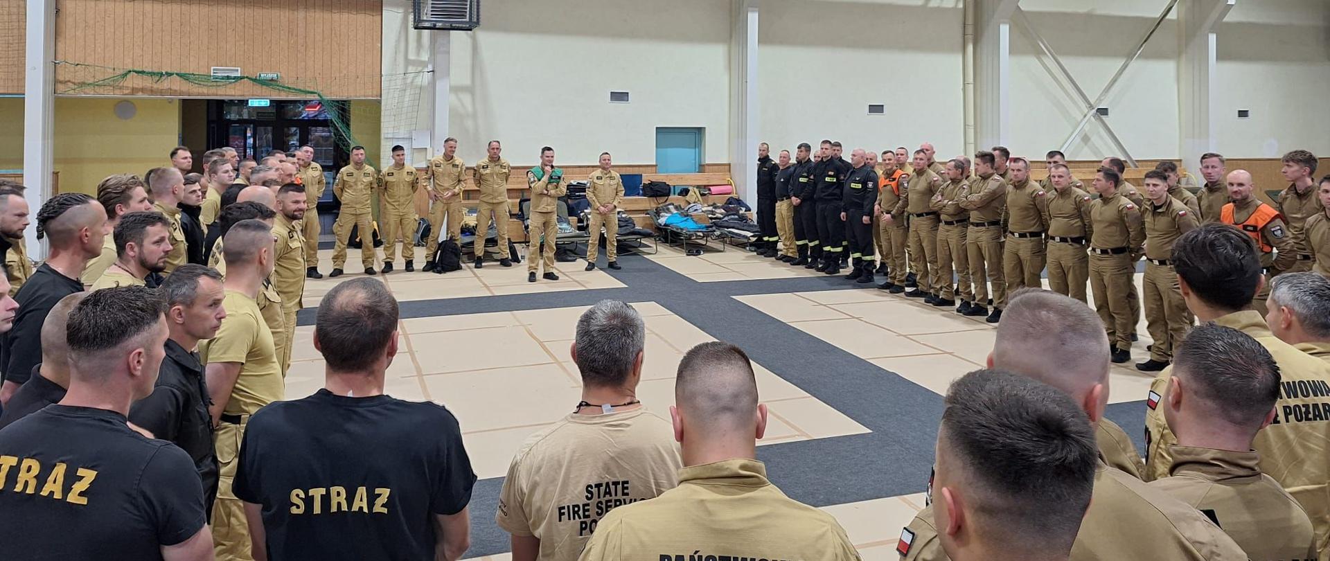 Duża grupa strażaków Państwowej Straży Pożarnej stoi w szerokim kręgu w jasnej hali treningowej, ubrana w beżowe mundury z napisami ‘State Fire Service Poland’. W tle widać sprzęt i torby ułożone pod ścianami