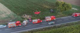 Na drodze krajowej duża ilość zastępów pożarniczych, karetka pogotowia oraz patrol Policji. Na pobliskim polu widoczny rozbity pojazd osobowy oraz parawan ratowniczy za którym działania medyczne prowadzą służby ratunkowe. Ruch pojazdów odbywa się wahadłowo. 