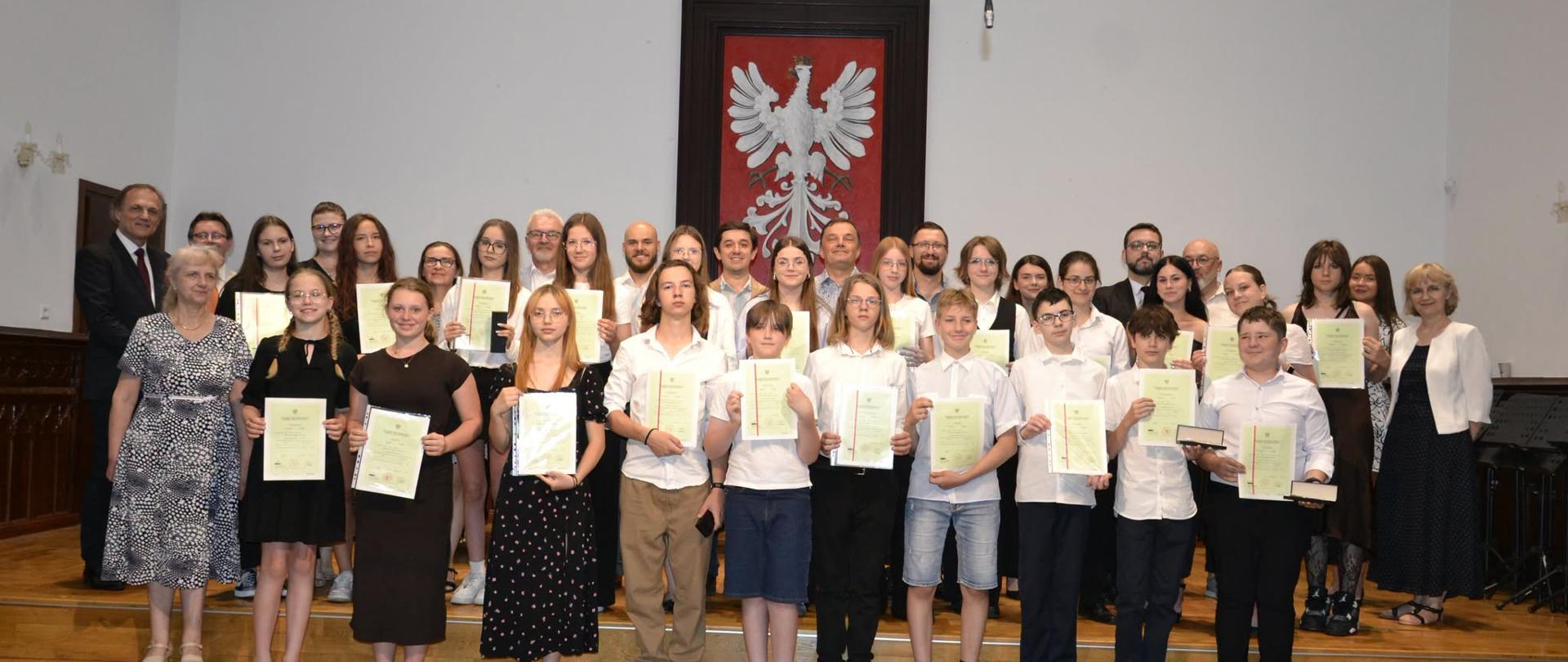 Grupa absolwentów PSM I stopnia trzymających świadectwa ukończenia szkoły pozuje wraz z nauczycielami na scenie Sali Królewskiej, w tle herb Polski i freski z portretami królów.