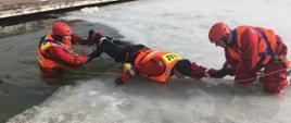 Na zdjęciu widać 3 funkcjonariuszy Państwowej Straży Pożarnej. Strażacy symulują wydobycie osoby pod którą zarwał się lód. Strażacy w umundurowaniu przeznaczonym do działań na wodzie i lodzie. 