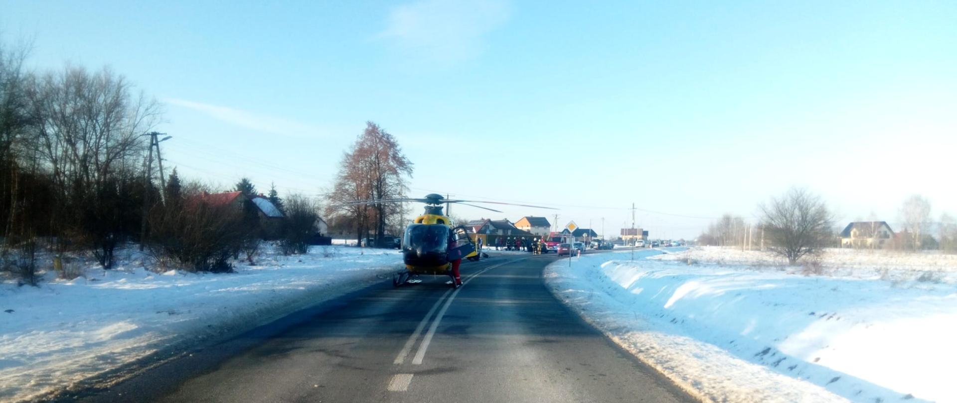 Zdjęcie przedstawia ulicę Częstochowską. Na pasie jezdni wylądował śmigłowiec. Na dalszym planie uszkodzone pojazdy, samochody służb ratowniczych oraz ratownicy.