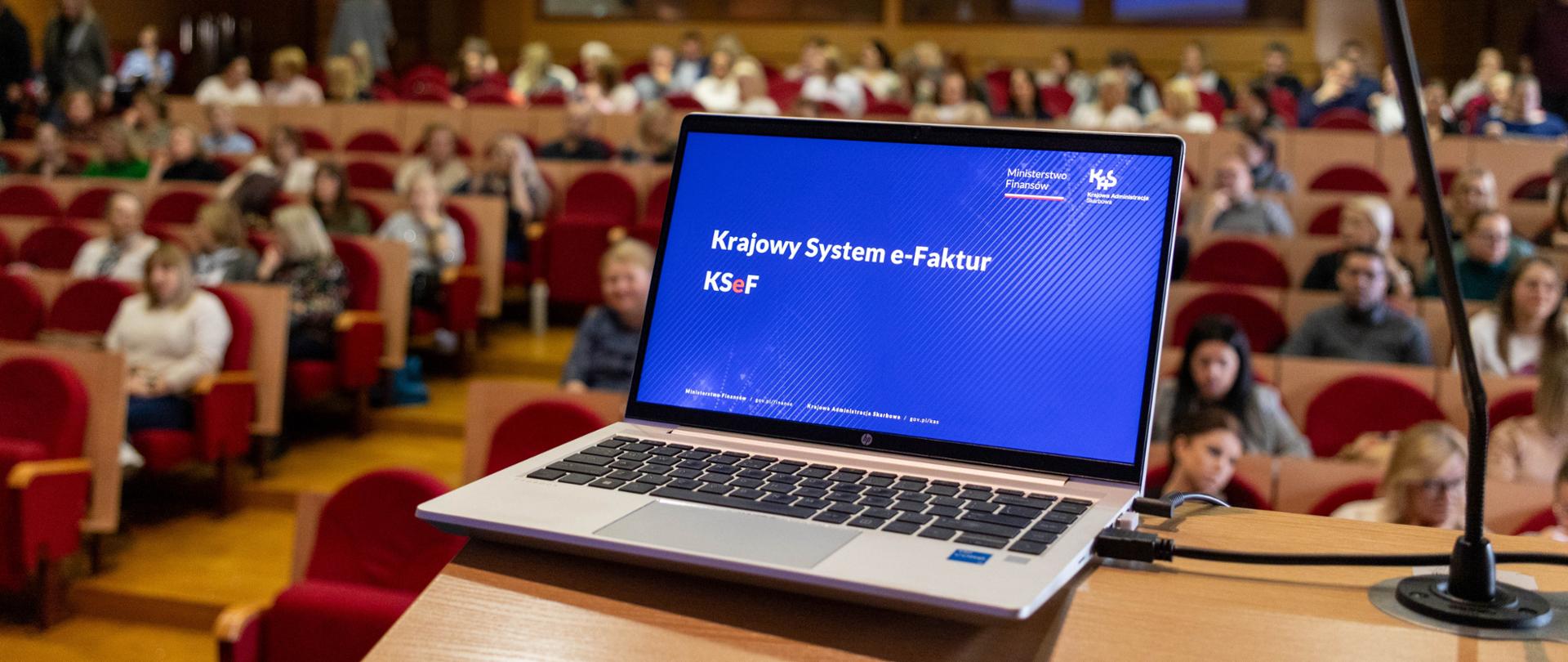 Laptop wyświetlający napis Krajowy System e-Faktur, w tle zebrani ludzie