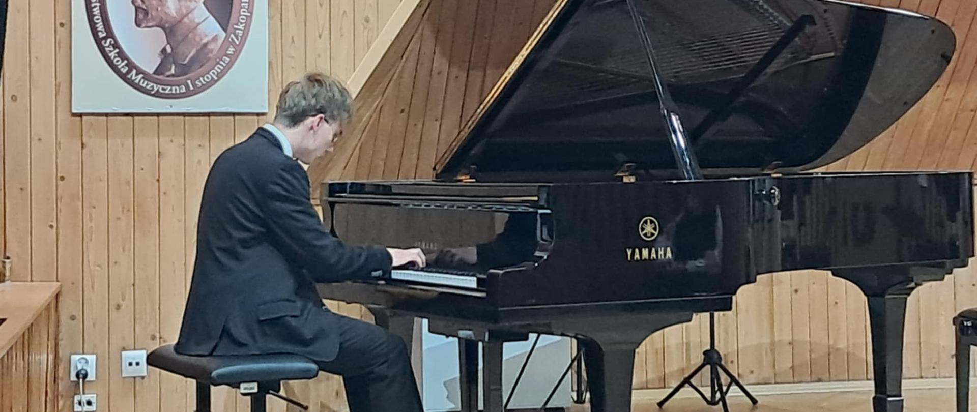 Zdjęcie przedstawia ucznia ubranego w garnitur, siedzącego tyłem do widza i grającego na czarnym fortepianie marki Yamaha. Fortepian jest otwarty, ukazując jego mechanizm i struny. Mężczyzna ma krótkie, jasne włosy. Scena rozgrywa się w pomieszczeniu o drewnianych, pionowych panelach na ścianach, które nadają jej ciepły i akustyczny wygląd. Po lewej stronie ściany, za pianistą, wisi duży portret starszego mężczyzny w profilu, otoczony napisem "Państwowa Szkoła Muzyczna I stopnia w Zakopanem imienia Mieczysława Karłowicza". Obok portretu, w dolnym lewym rogu zdjęcia, widać fragment jasnego, prostokątnego obiektu, prawdopodobnie części innej ściany lub mebla. Oświetlenie w pomieszczeniu jest rozproszone, ale dobrze eksponuje fortepian i grającego na nim pianistę. Całość kompozycji zdjęcia jest spokojna i skupiona na akcie muzykowania.