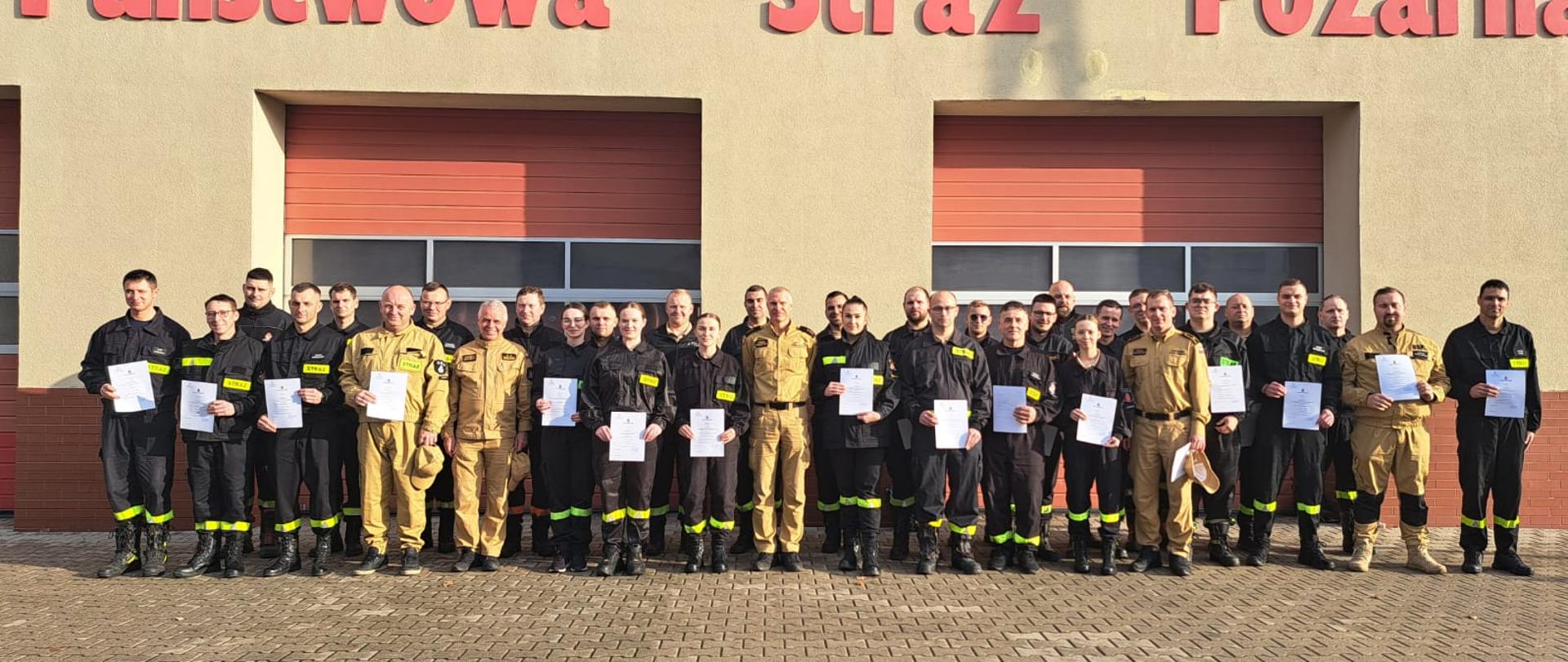 Uczestnicy szkolenia na kierującego działaniem ratowniczym dla strażaków ratowników ochotniczych straży pożarnych stoją przed budynkiem KP PSP w Rawiczu. Przed sobą trzymają zaświadczenia, świadczące o pozytywnym ukończeniu szkolenia. Pomiędzy nimi strażacy z KP PSP w Rawiczu, którzy prowadzili szkolenie.