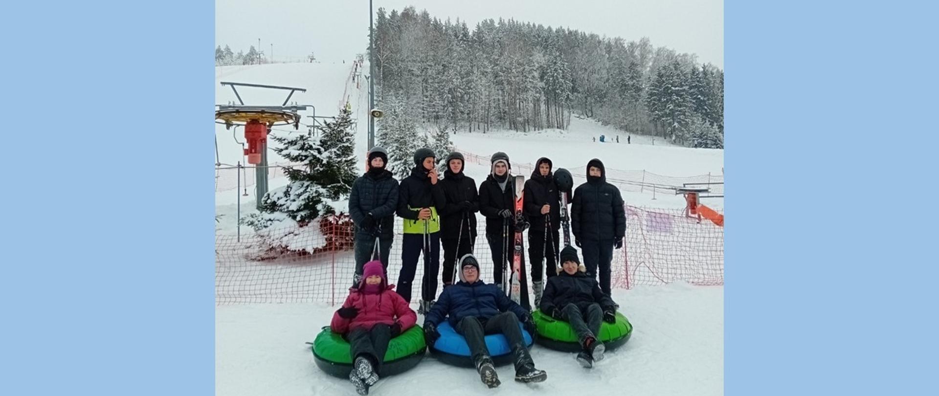 Grupa młodych osób pozuje na ośnieżonym stoku narciarskim, stojąc z nartami lub siedząc na kolorowych pontonach do snowtubingu. W tle rozpościera się wyciąg narciarski oraz las pokryty szronem pod zachmurzonym niebem.