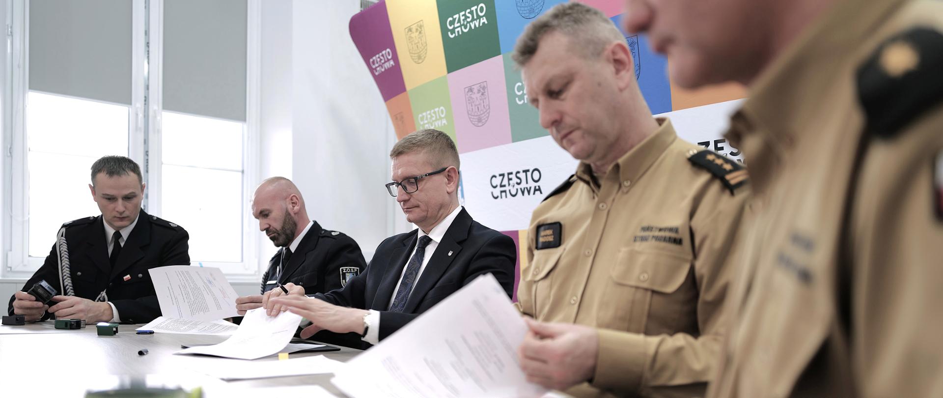 Na zdjęciu widać kilku mężczyzn siedzących przy stole podczas oficjalnego spotkania. Część z nich ma na sobie mundury służbowe (ciemne i piaskowe), a jeden z mężczyzn jest ubrany w garnitur. Wszyscy przeglądają i podpisują dokumenty. W tle znajduje się kolorowa ścianka z herbem i napisem „Częstochowa”.