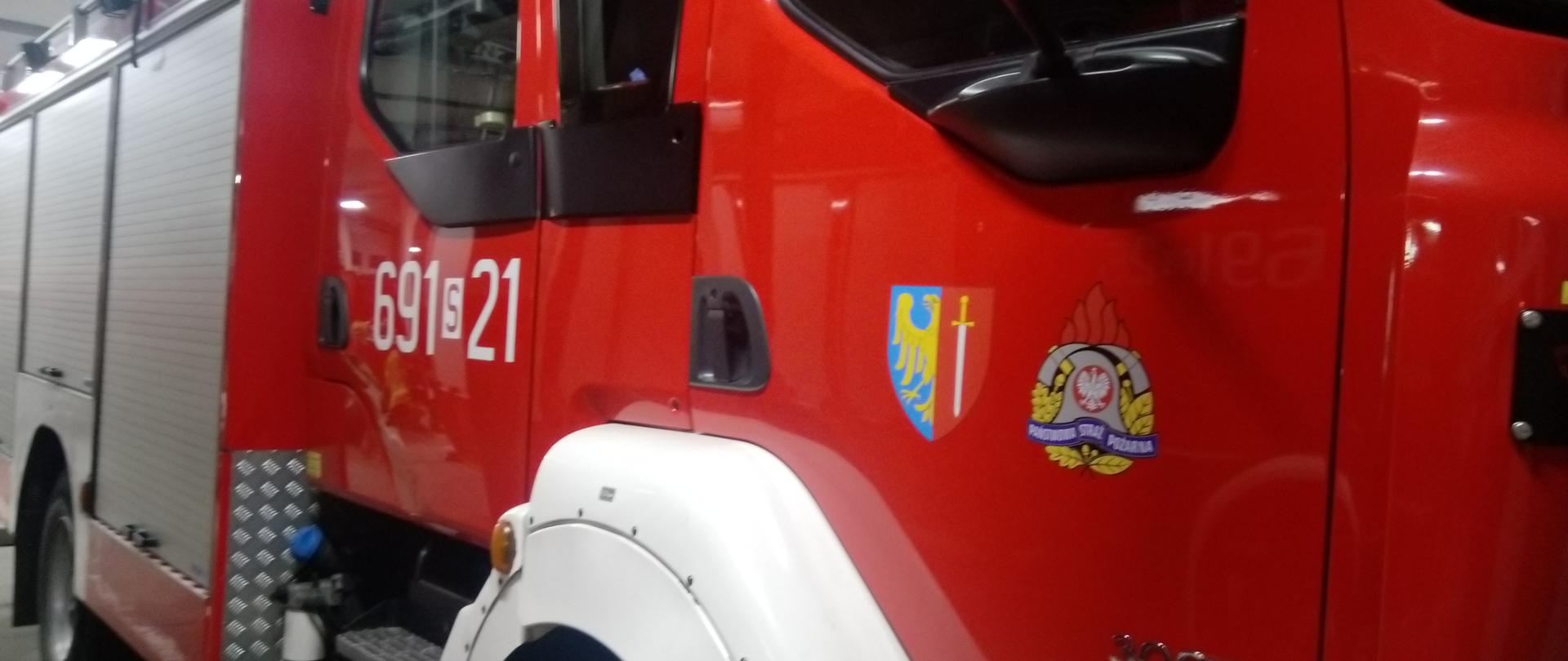 Czerwony wóz strażacki, na pierwszym planie prawe drzwi samochodu z logo Państwowej Straży Pożarnej oraz Miasta Żory dalej żaluzje skrytek sprzętowych samochodu