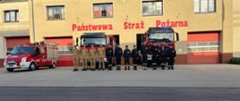 Plac przed komendą. Na tle budynku komendy i wozów bojowych, w szeregu stoją strażacy PSP i OSP, którzy oddają hołd tragicznie zmarłemu strażakowi OSP. Dowódca stojący na prawym skrzydle oddaje honor przez salutowanie. Czterech strażaków ma na sobie ubranie służbowe w kolorze piaskowym, pozostali w ciemnym.
