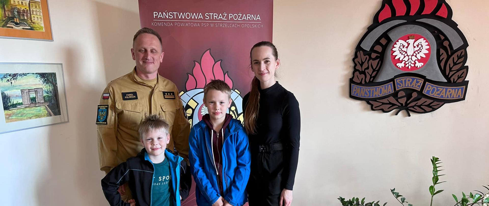 Na zdjęciu znajduje się dwoje strażaków w ubraniach służbowych oraz dwoje dzieci. Ustawieni są w gabinecie, na ścianach znajdują się baner oraz logo PSP