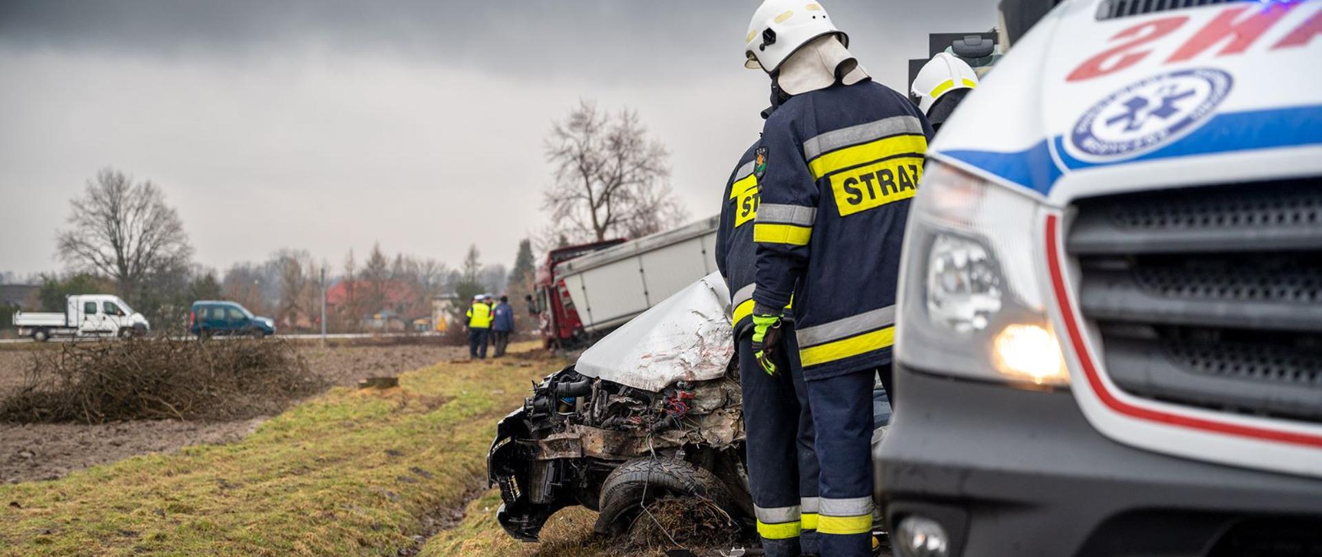 Na zdjęciu widoczny uszkodzony samochód osobowy w tyle samochód ciężarowy w rowie obok karetka ZRM