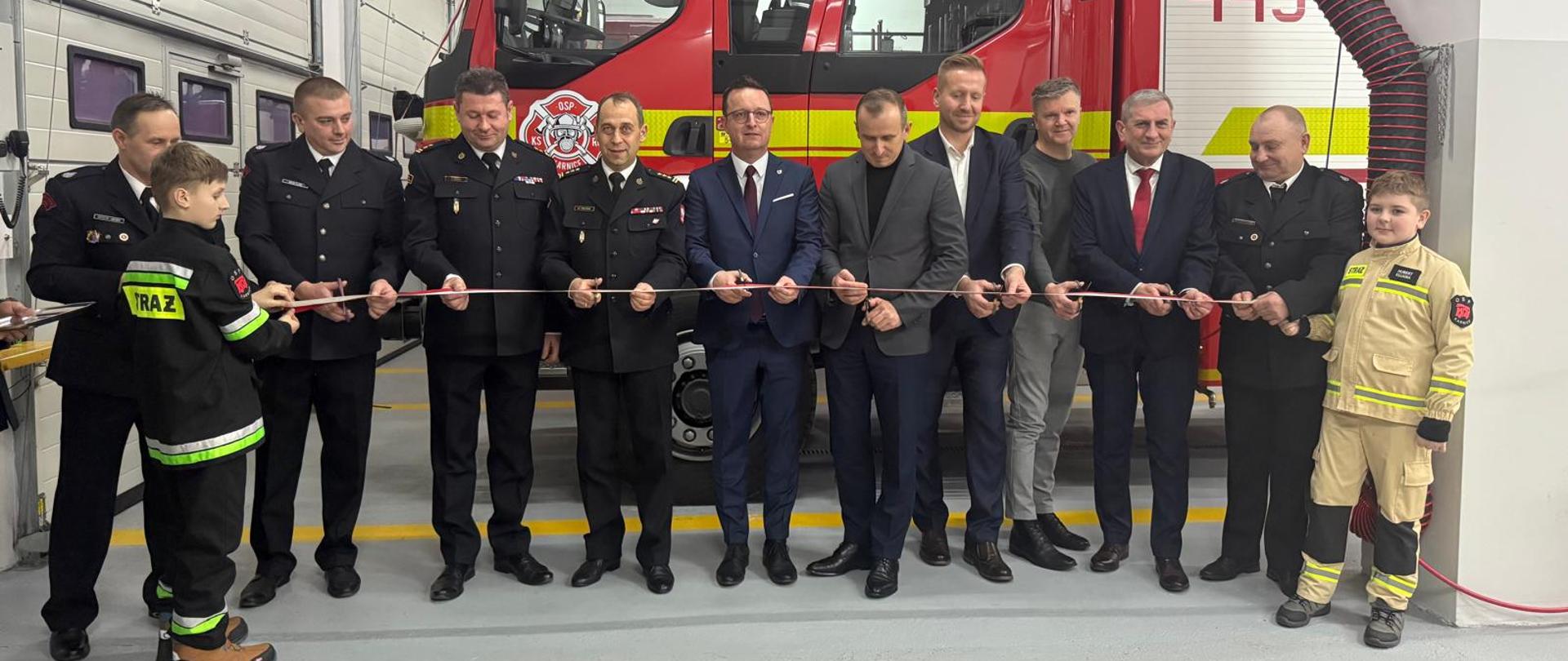 zachodniopomorski komendant wojewódzki państwowej straży pożarnej, w towarzystwie zaproszonych gości, podczas uroczystości przekazania nowego samochodu ratowniczo-gaśniczego dla ochotniczej straży pożarnej w karnicach. W tle widoczny jest nowy pojazd jednostki osp. zdjęcie powiększy się po kliknięciu na nie.