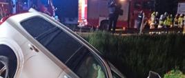 Zdjęcie przedstawia biały, zniszczony samochód osobowy znajdujący sie w rowie. Nad samochodem stoją strażacy oraz wozy strażackie