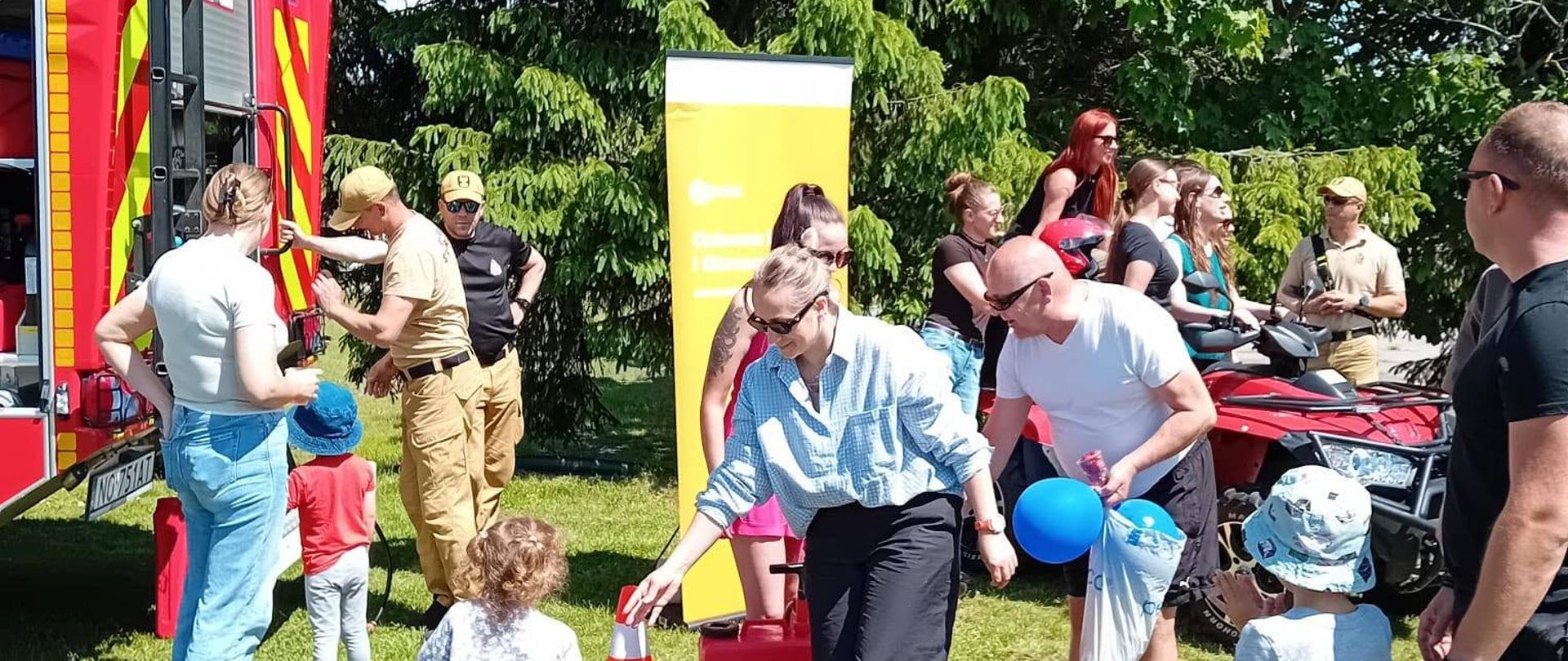 Na zdjęciu zabawa dzieci z rodzicami przy sprzęcie strażackim. Zielony park, słonecznie.