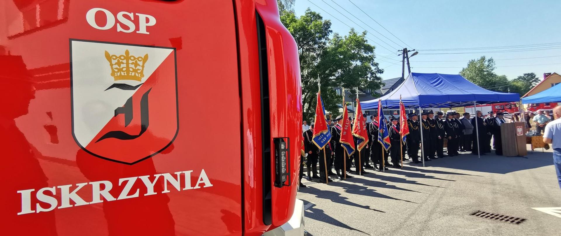 Na pierwszym planie przód samochodu strażackiego. W tle ustawione poczty sztandarowe.