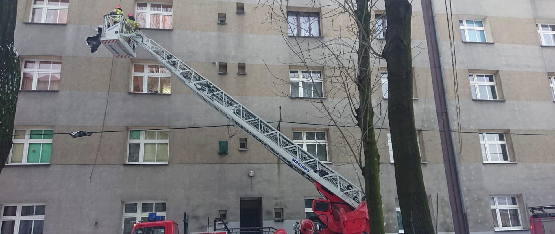 Zdjęcie przedstawia samochód pożarniczy z drabiną mechaniczną na tle budynku oraz strażaków usuwających nadpaloną elewację budynku 