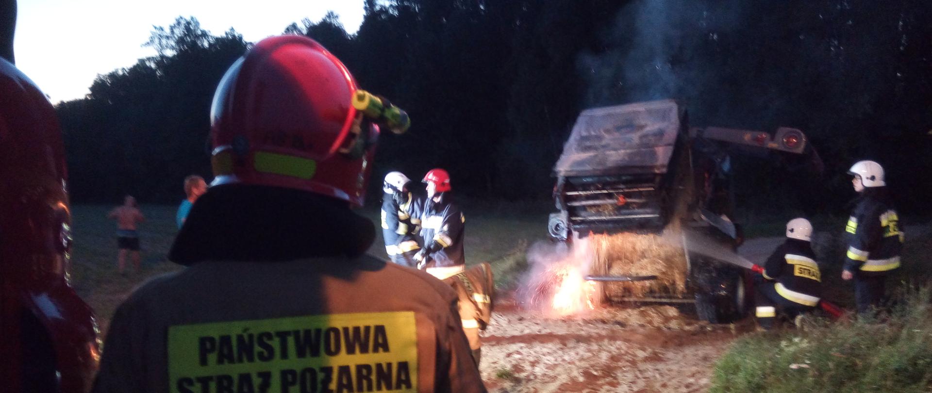 Strażacy podczas gaszenia prasy belującej. Na maszynie widoczne są ślady opalenia. Nad belarką unosi się dym.