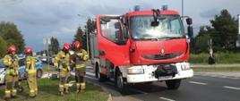 Samochód strażacki na poboczu drogi. Obok strażacy uczestniczący w działaniach. W tle samochód policyjny oraz inne samochody czekające na możliwość przejazdu.