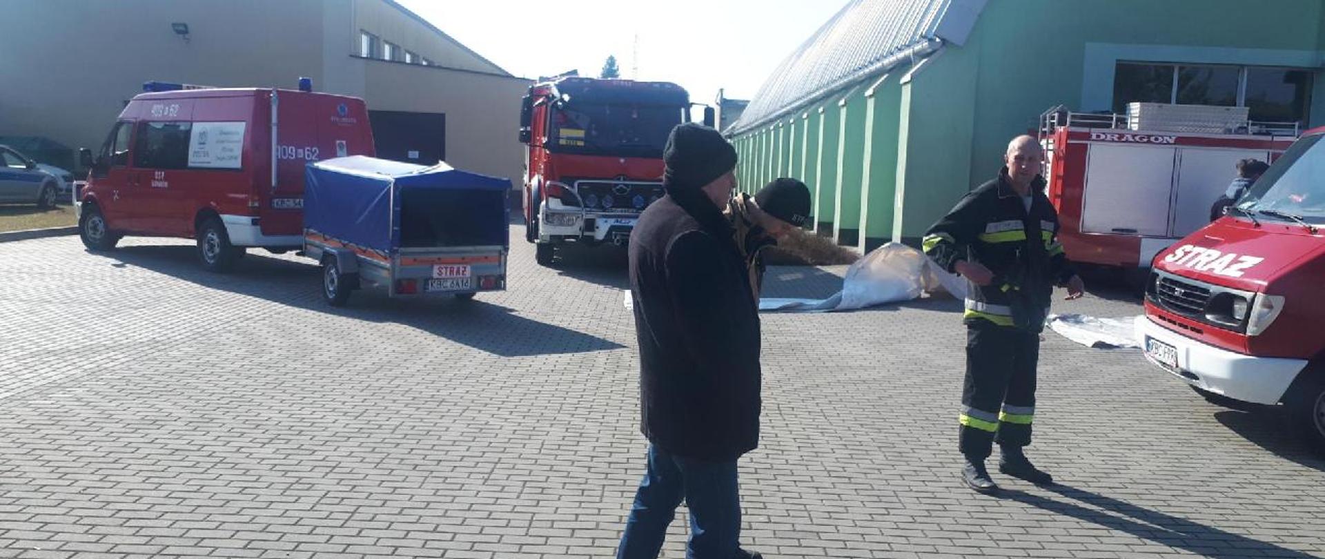 Na pierwszym planie strażacy w mundurach pracują przy rozładunku namiotów, w których będą gromadzone dary dla uchodźców z Ukrainy. Na drugim planie stoją 4 samochody strażackie i jedna przyczepka z namiotami