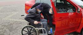Strażak pomaga zsiąść z wózka inwalidzkiego starszemu mężczyźnie i przesiąść się do samochodu strażackiego, którym uda się na szczepienie