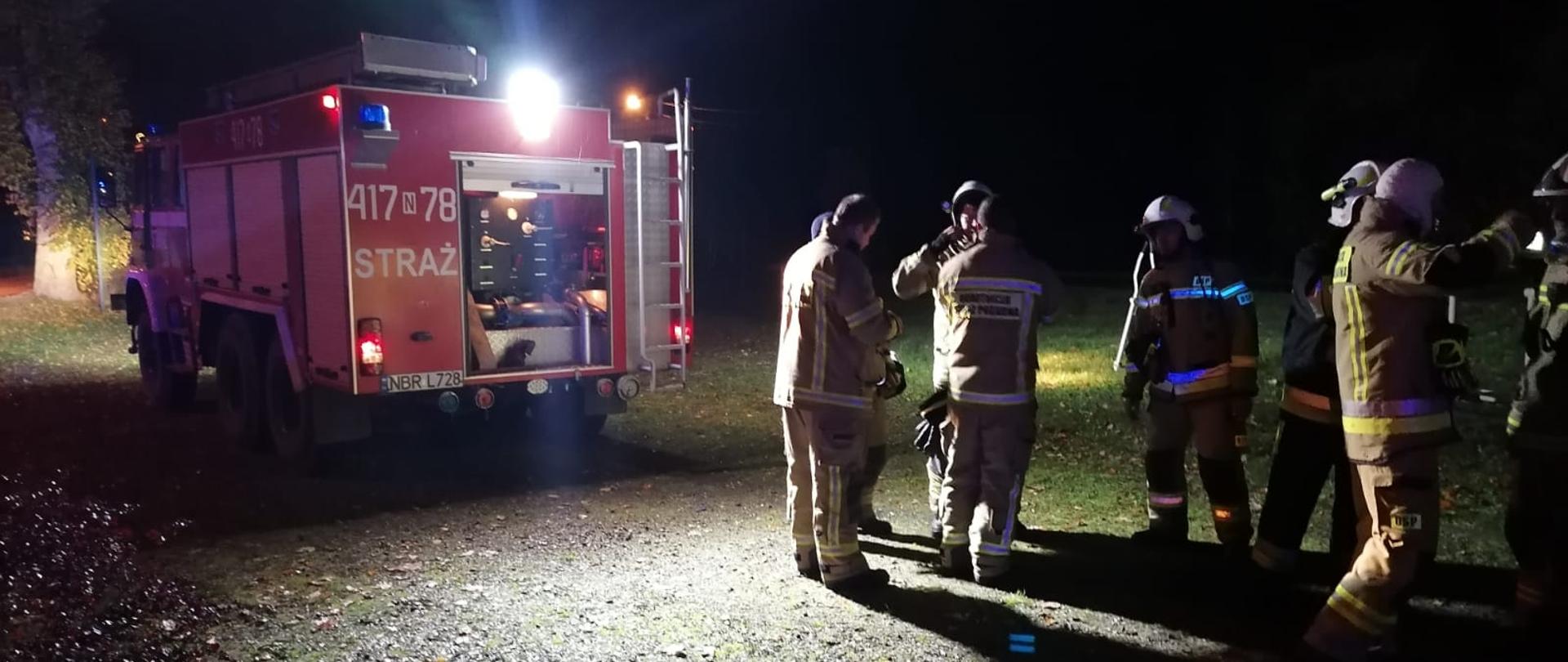 Zdjęcie zrobione nocą. Z lewej strony stoi samochód strażacki na światłach. Z prawej kilku strażaków.